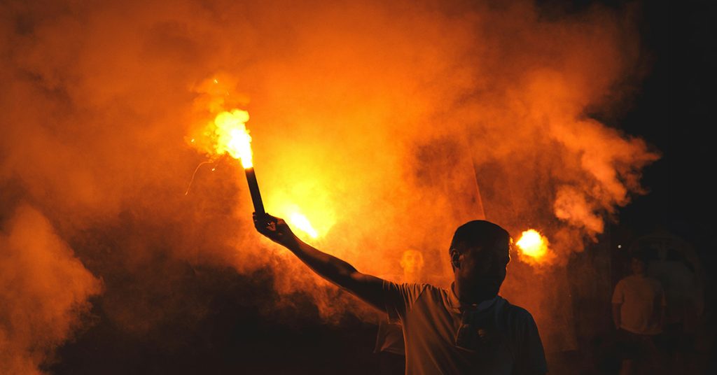Ein Mann hält bei Nacht eine brennende Signalfackel in die Höhe, umgeben von orangerotem Rauch.